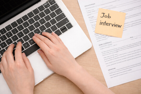 Hände auf Laptop-Tastatur neben Bewerbungsunterlagen und Notizzettel „Job interview“